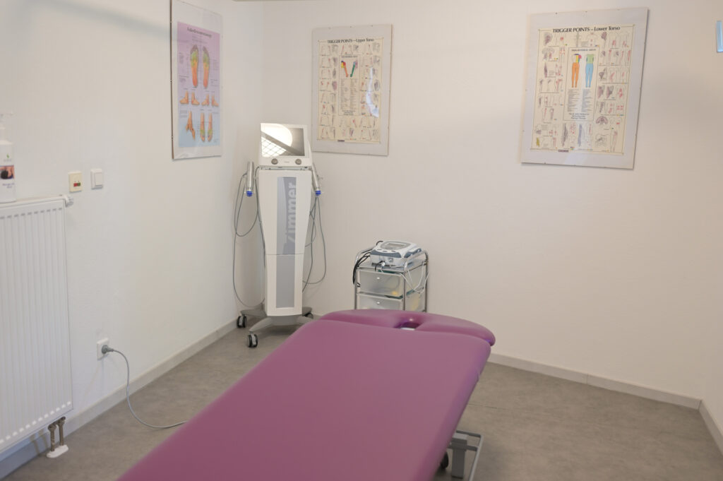 Ein heller Behandlungsraum mit einer lila Liege und medizinischen Geräten.