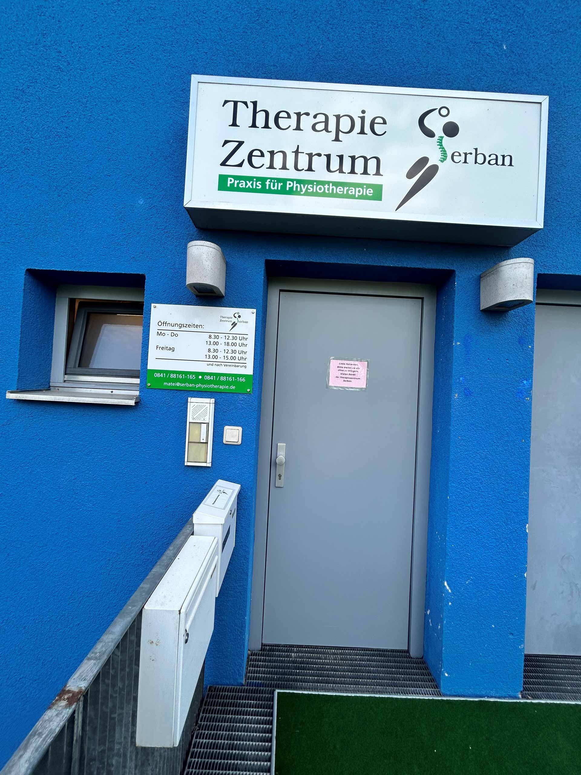 Blauer Gebäudeeingang mit Schild "Therapie Zentrum", daneben eine graue Tür.