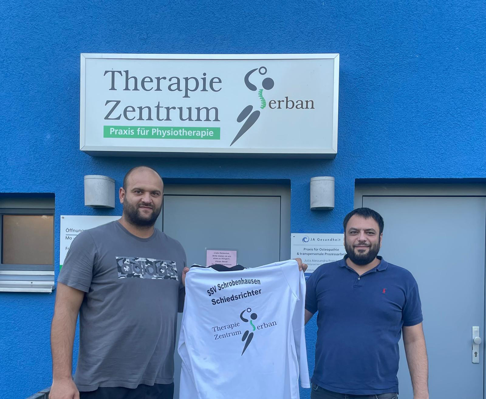 Zwei Männer stehen vor einem Therapiezentrum und halten ein Trikot.